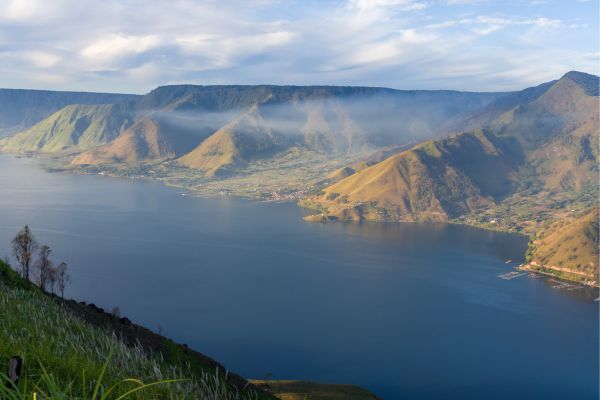 Wide view of Lake Toba surrounded by rolling green hills and misty mountain ridges under a cloudy sky.