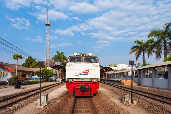 White and red Indonesian train stopped at a small tropical station with palm trees and a blue sky.